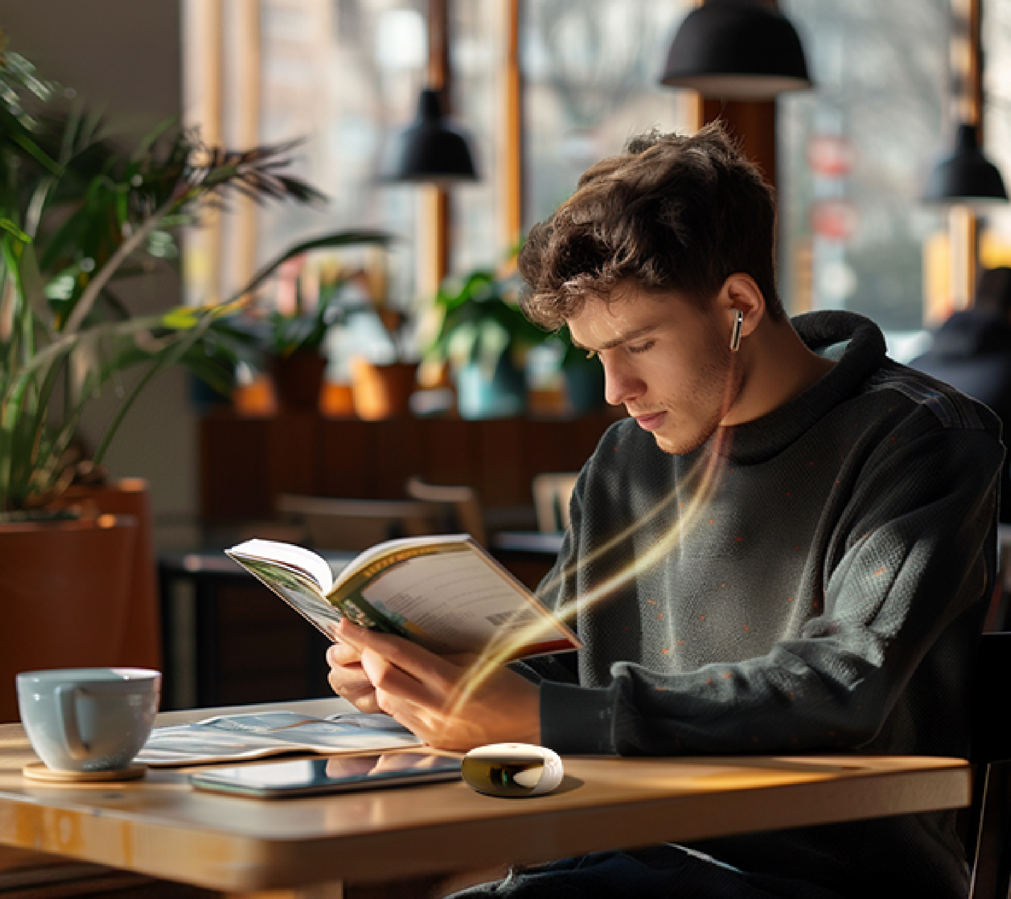 Jeune homme lisant un livre dans un café avec des écouteurs sans fil et une tasse de café sur la table en bois.
