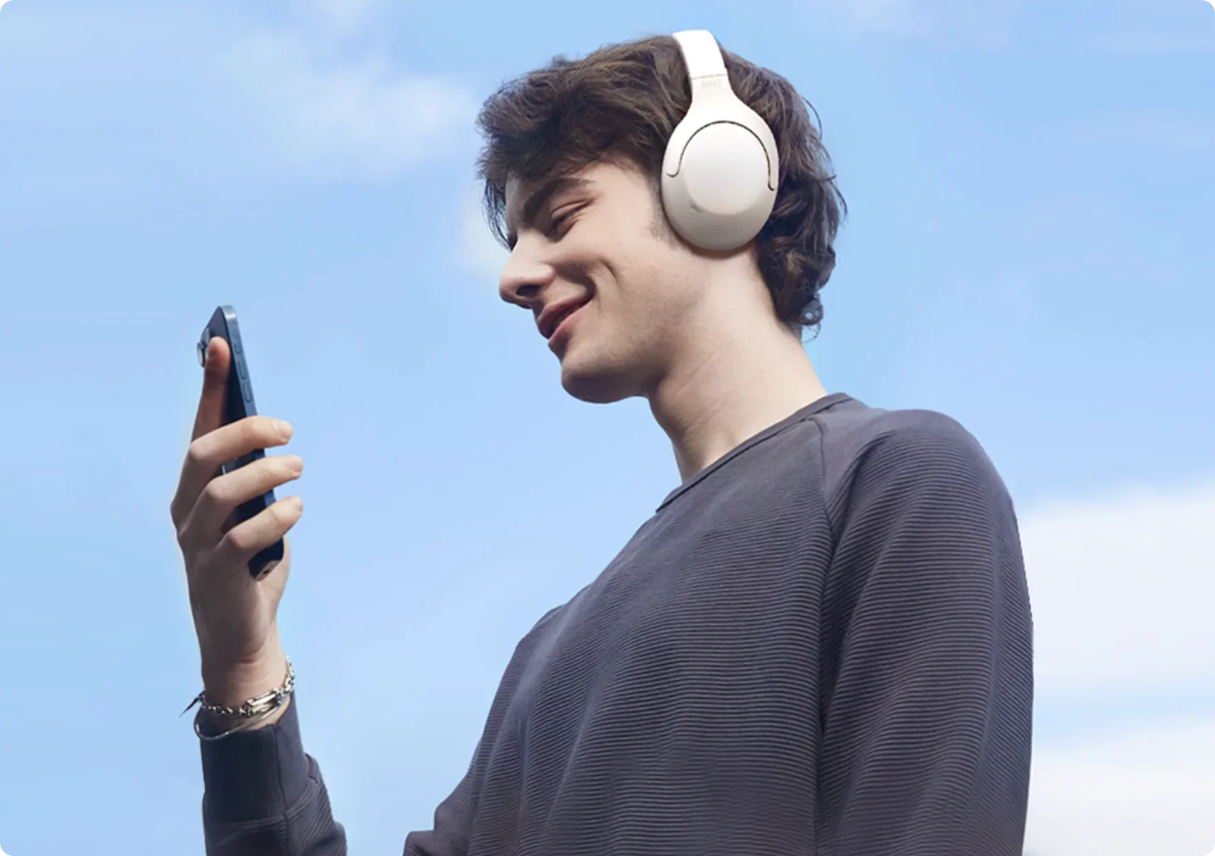 Jeune homme avec des écouteurs blancs et un smartphone sous un ciel bleu.
