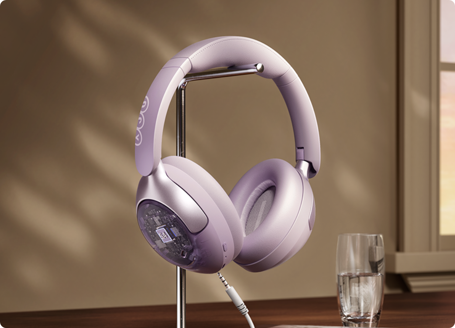 Casque circum-auriculaire lavande avec boîtier transparent sur support, câble branché, à côté d'un verre d'eau près de la fenêtre.
